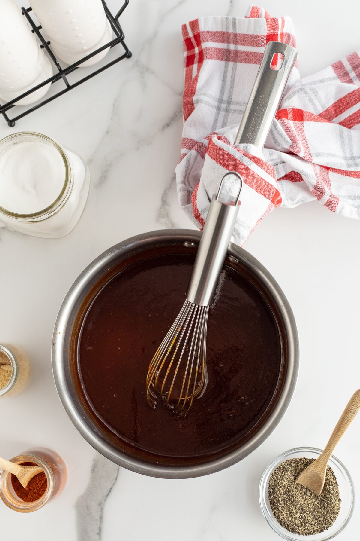 A metal bowl filled with dark barbecue sauce and a whisk, surrounded by jars of spices, a bowl of pepper, and a red-and-white kitchen towel on a white marble surface.