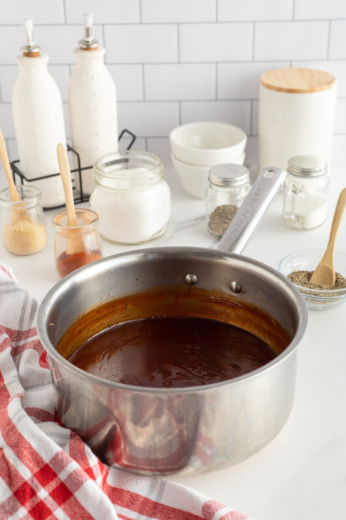 A stainless steel saucepan filled with dark brown sauce sits on a white countertop. Surrounding it are jars of seasonings, a bowl, two white bottles, and a red-and-white checkered towel.