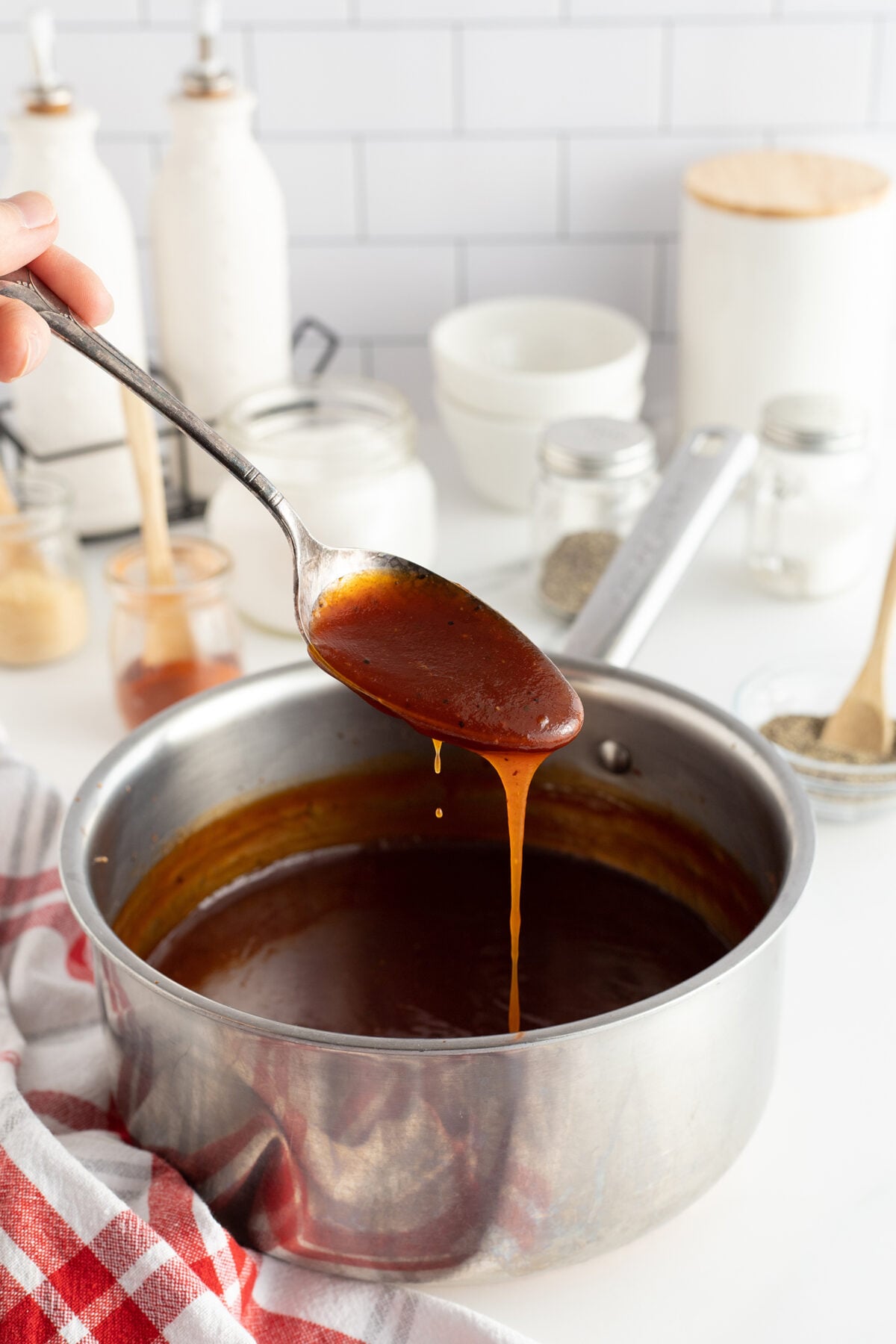 A spoon holds up dripping barbecue sauce over a saucepan, with various jars and kitchen utensils on a white countertop in the background.