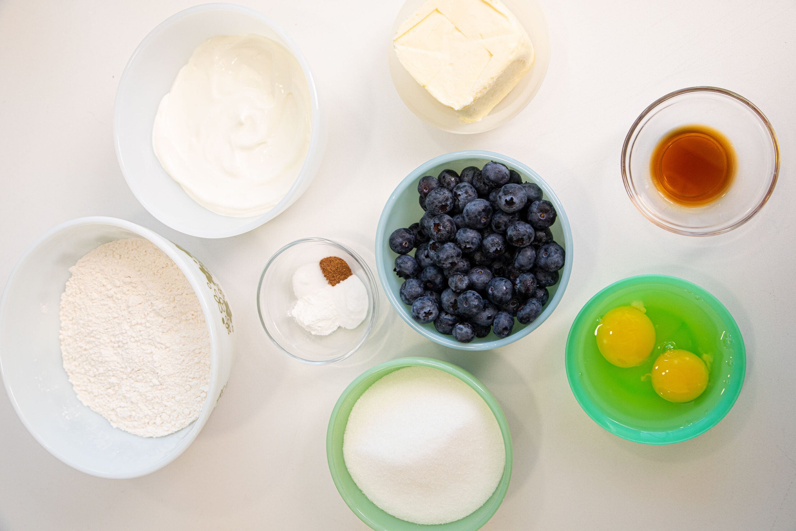 Bowls with baking ingredients on a white surface: flour, sugar, butter, sour cream, two eggs, blueberries, baking powder with spice, and vanilla extract.