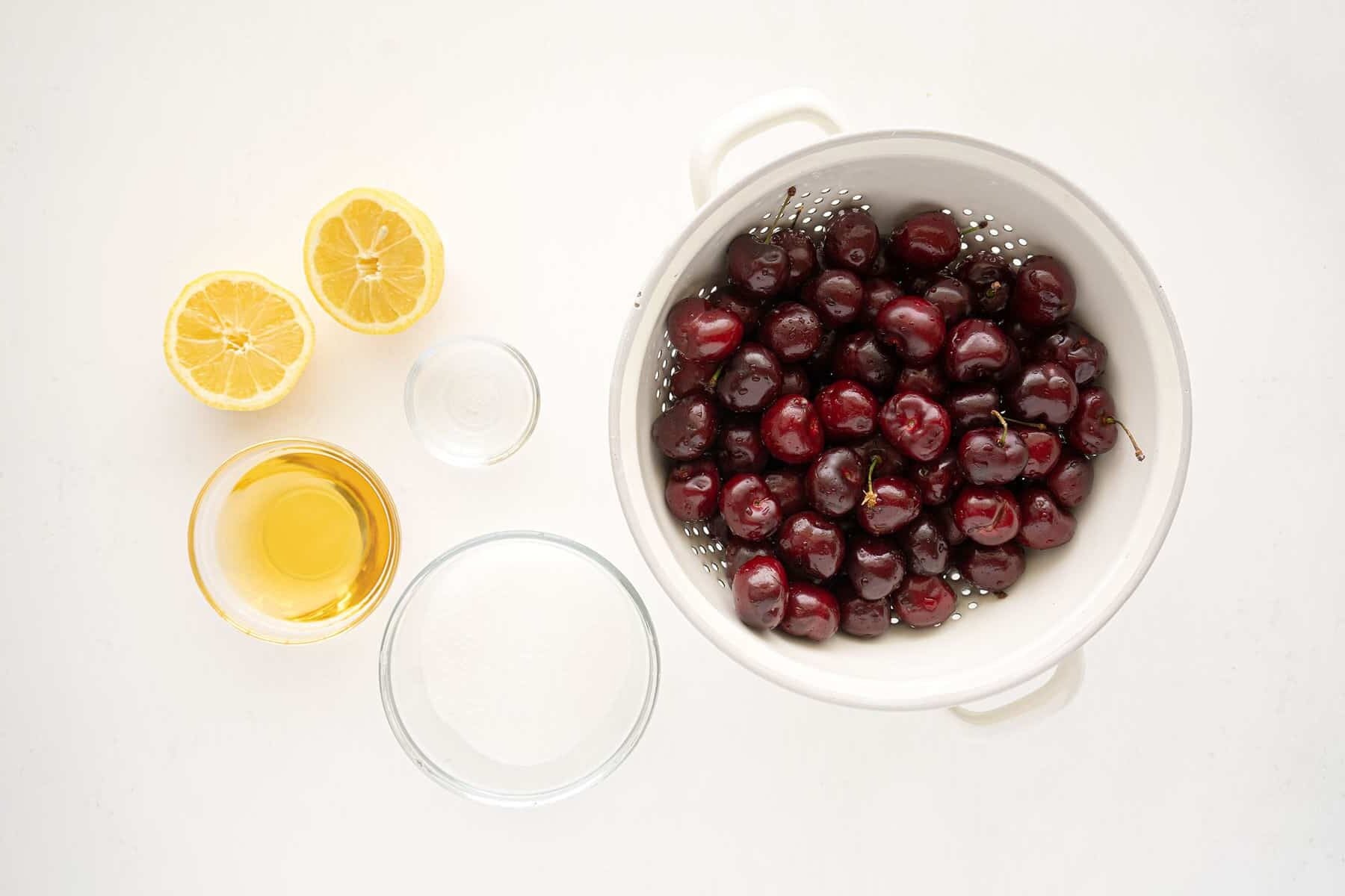 A white colander filled with fresh cherries sits next to two lemon halves, a small bowl of sugar, a bowl of honey, and a small dish of clear liquid on a white surface.