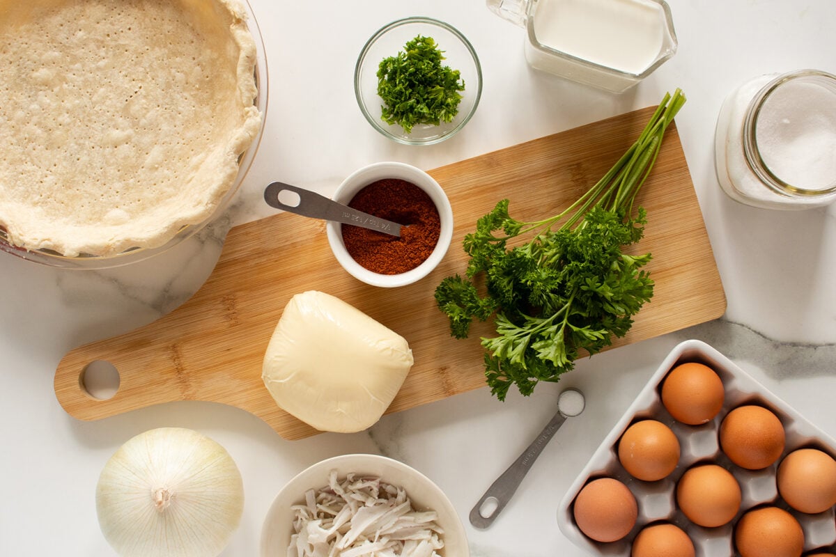 A top view of ingredients on a white surface: a pie crust, block of butter, fresh parsley, a bowl of shredded chicken, white onion, eggs, paprika, chopped parsley, milk, and a jar of sugar.