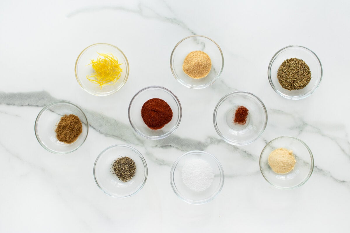 Nine small glass bowls filled with various spices, herbs, and seasonings are arranged neatly on a white marble surface, including yellow lemon zest, ground spices, salt, and pepper.