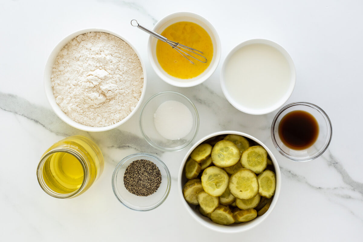 Bowls with flour, melted butter, milk, sugar, pickles, black pepper, and vanilla extract are arranged on a white marble surface. A jar of oil and a small whisk are also included.