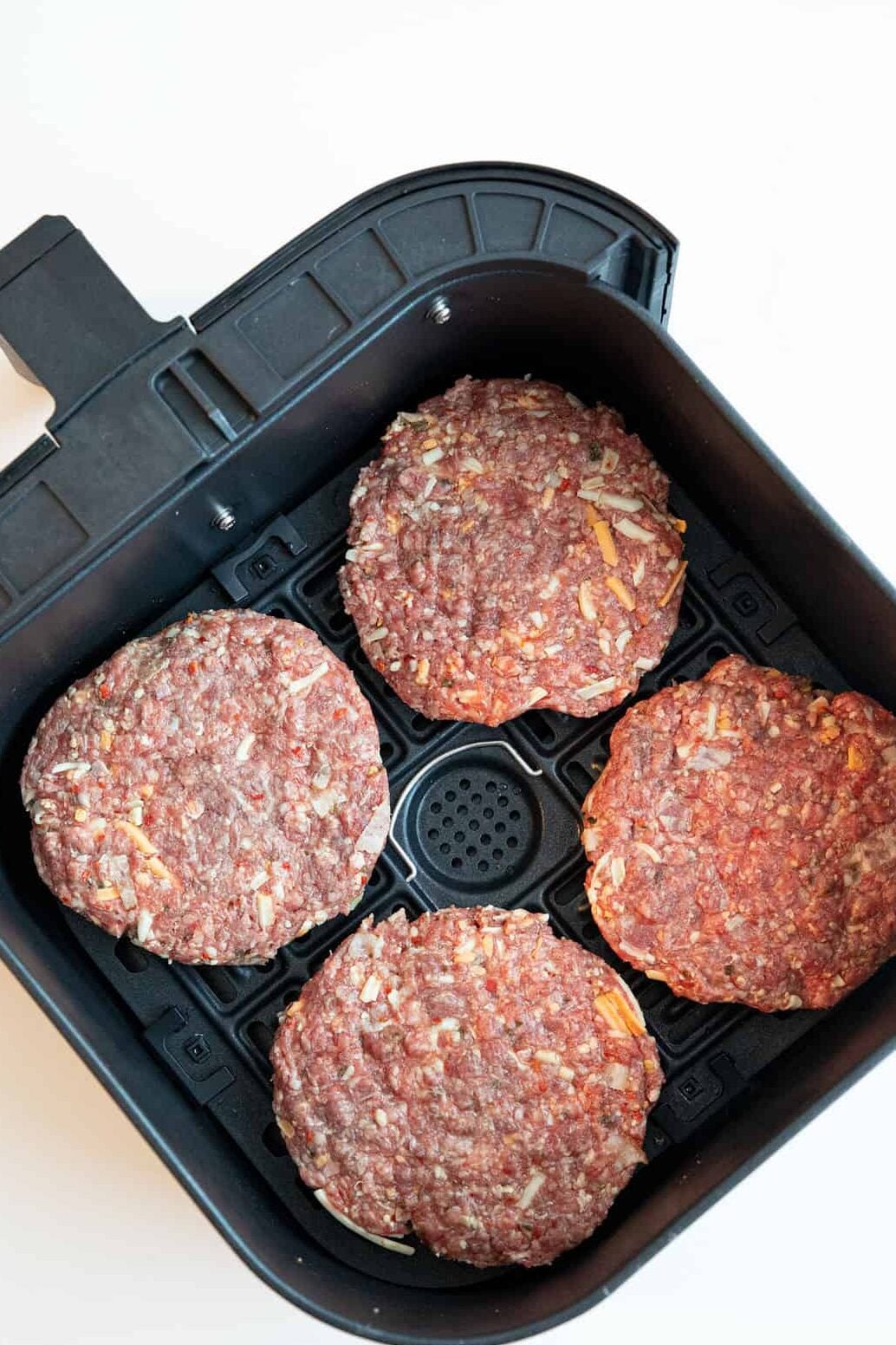 Four raw hamburger patties with visible onion and seasoning are arranged side by side in the basket of a black air fryer, ready to be cooked. The background is a plain white surface.