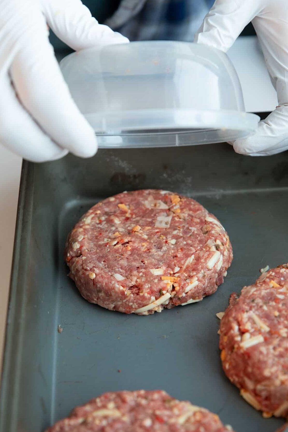 A person wearing white gloves uses a plastic container to shape raw ground meat patties with bits of cheese on a metal baking tray.