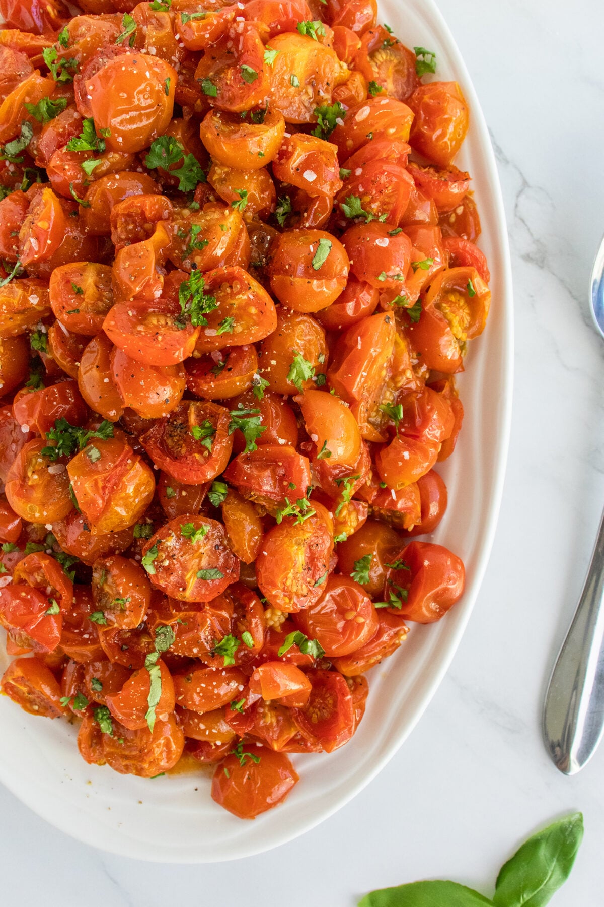 A white oval dish filled with roasted cherry tomatoes garnished with chopped herbs, placed on a marble surface with a spoon and fresh basil nearby.