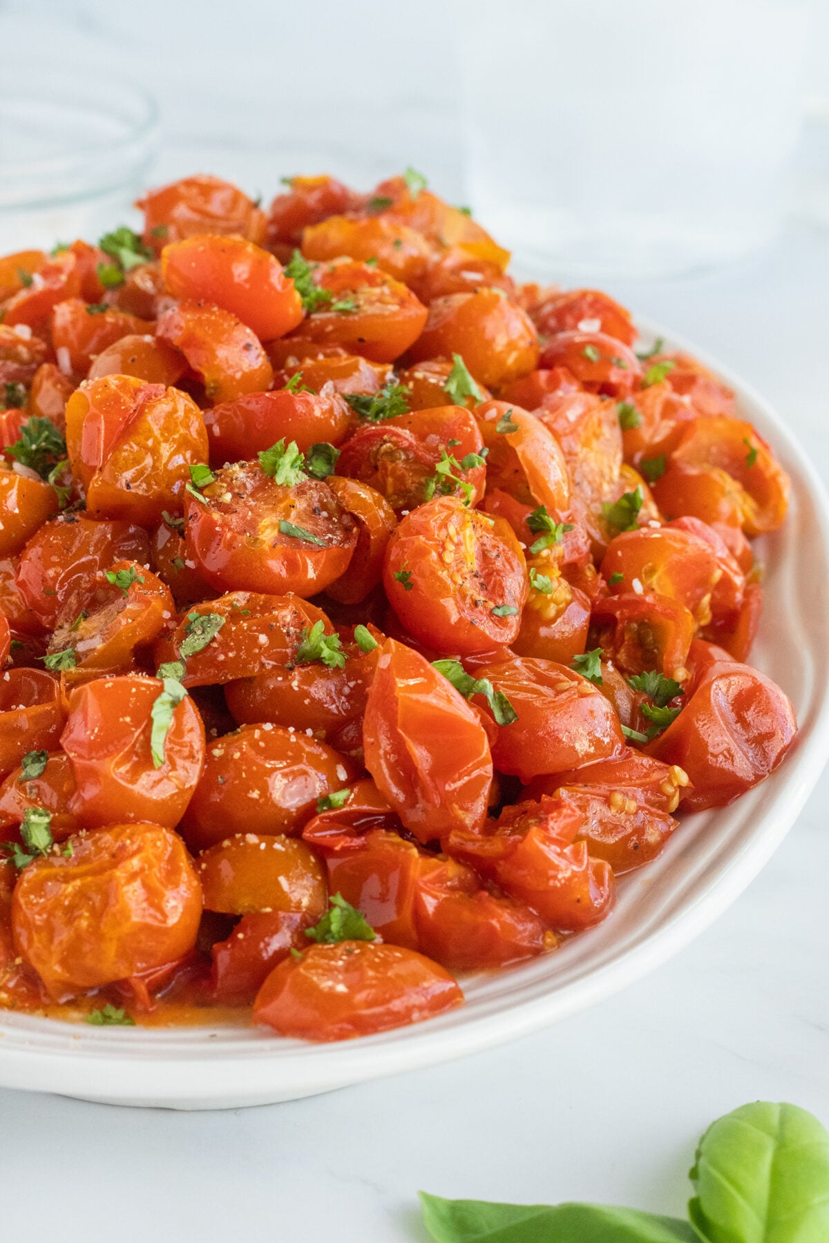 A white plate filled with roasted cherry tomatoes, garnished with chopped fresh herbs and a sprinkle of grated cheese, sits on a white surface with a basil leaf nearby.