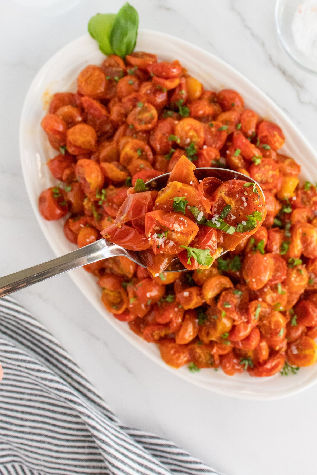 A serving spoon lifts a portion of roasted cherry tomatoes garnished with chopped basil from a white oval dish, with more tomatoes and basil visible on the plate below.