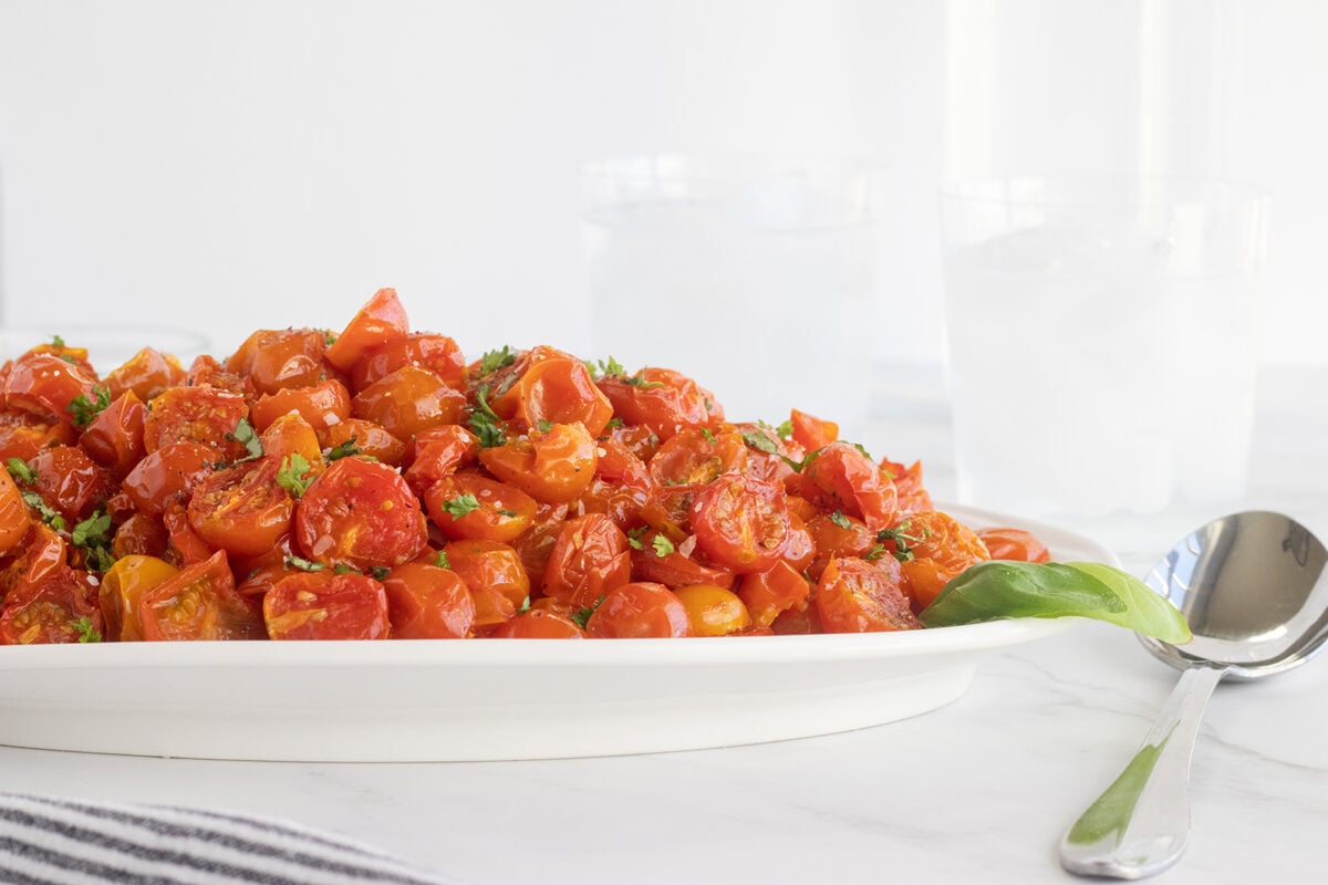 A white platter filled with roasted cherry tomatoes garnished with chopped herbs, set on a white marble surface beside a spoon and two glasses of ice water. A striped cloth is partially visible in the foreground.