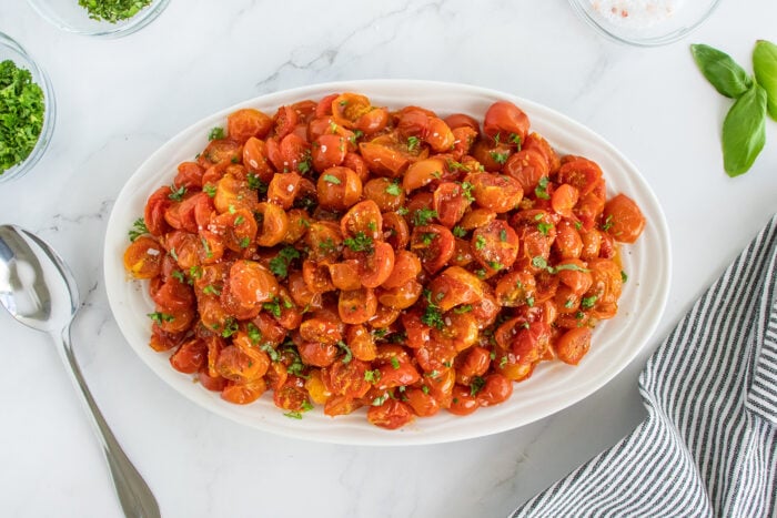 An oval white platter filled with a chopped cherry tomato salad, garnished with herbs. The platter is on a marble surface, with a spoon, a striped cloth, and bowls of herbs and salt nearby.