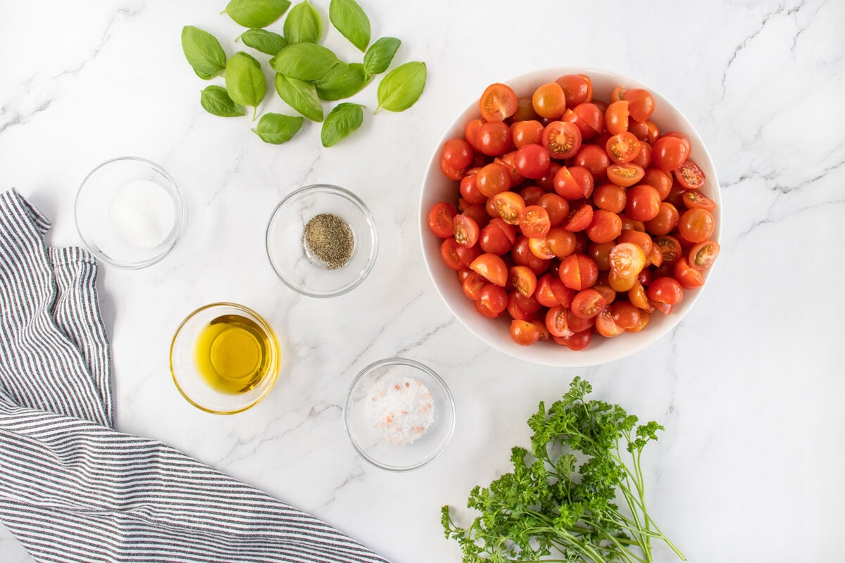 A bowl of halved cherry tomatoes, fresh basil leaves, parsley, bowls of olive oil, salt, and pepper are arranged on a white marble surface with a striped cloth on the side.