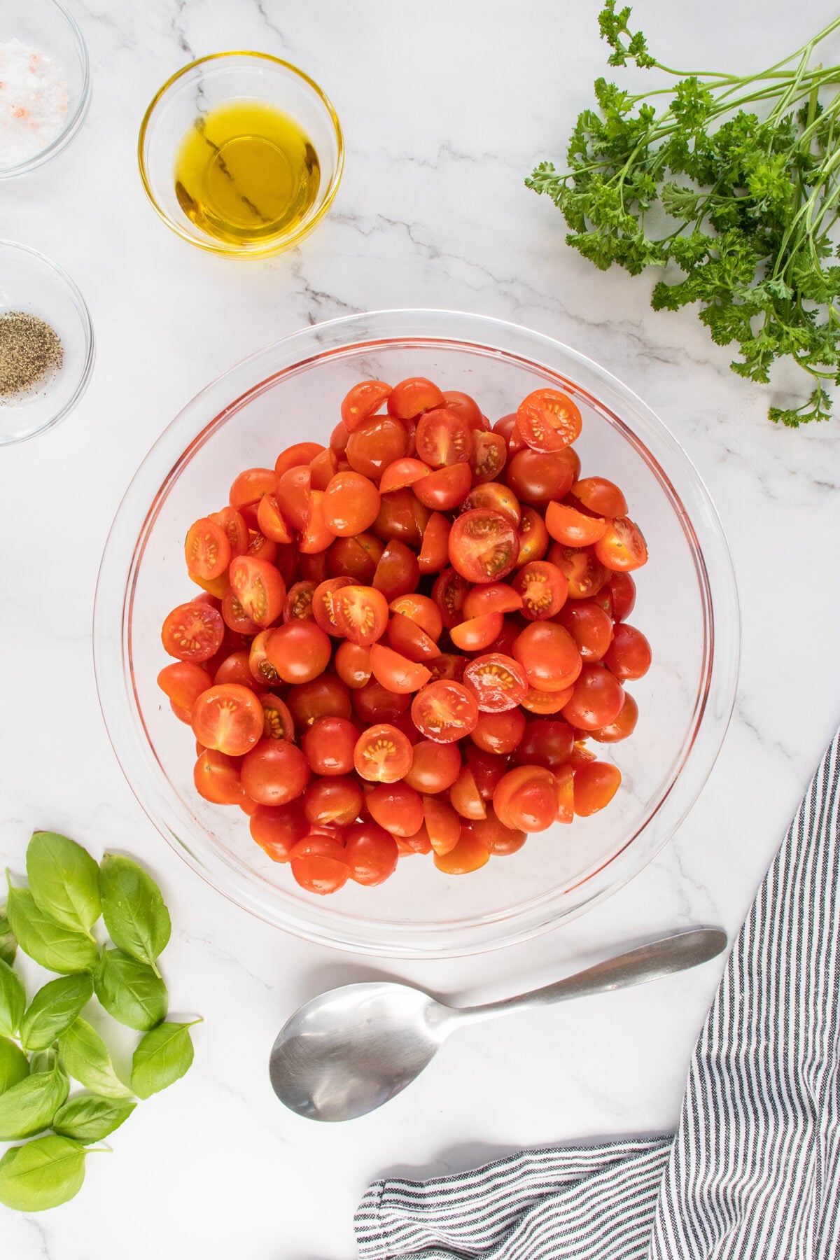 A glass bowl filled with halved cherry tomatoes sits on a marble countertop, surrounded by fresh basil, parsley, a bowl of olive oil, salt and pepper, and a striped kitchen towel with a silver spoon nearby.