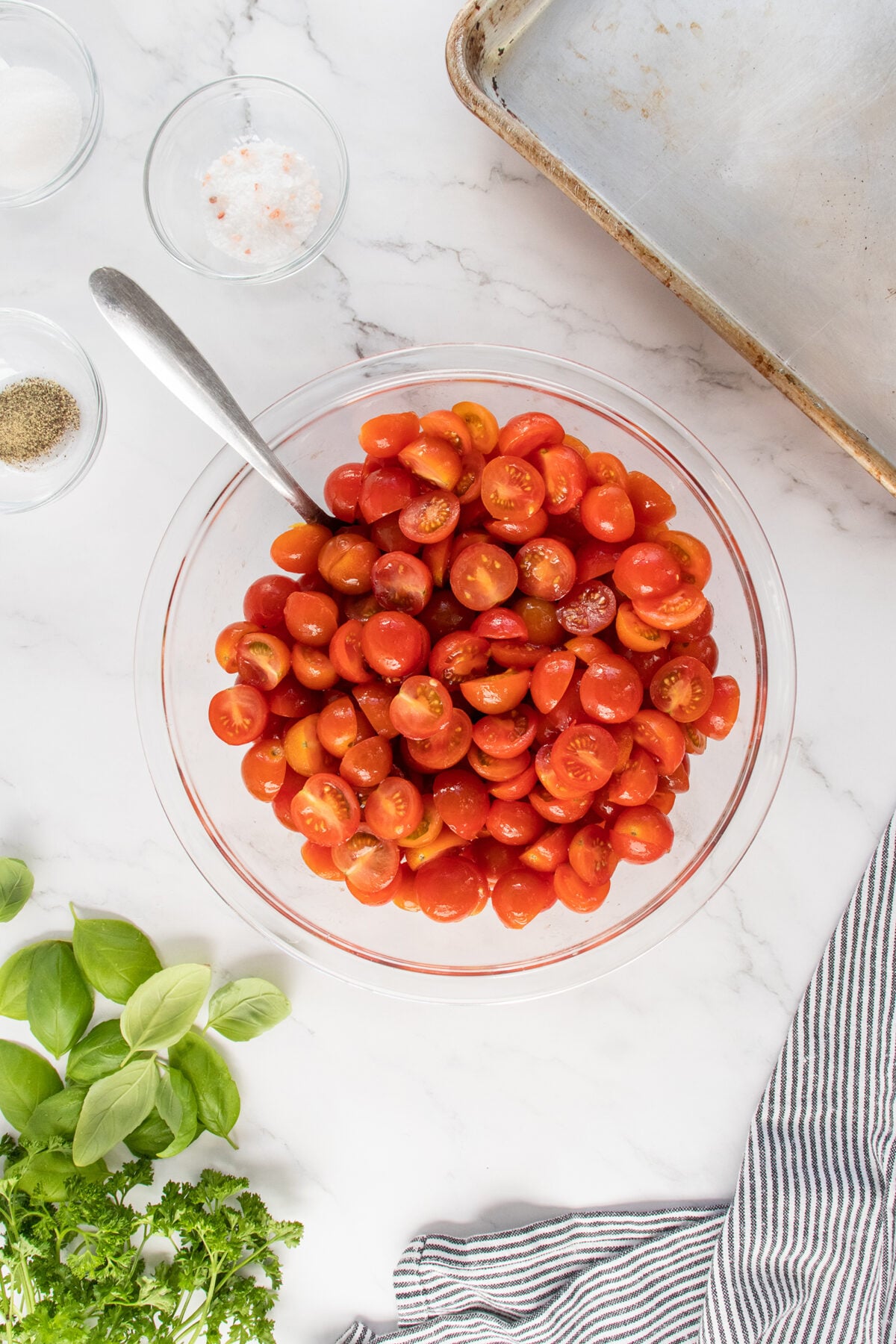 A glass bowl filled with halved cherry tomatoes and a spoon sits on a marble countertop, surrounded by fresh basil, parsley, small bowls of salt and pepper, a baking sheet, and a striped cloth napkin.