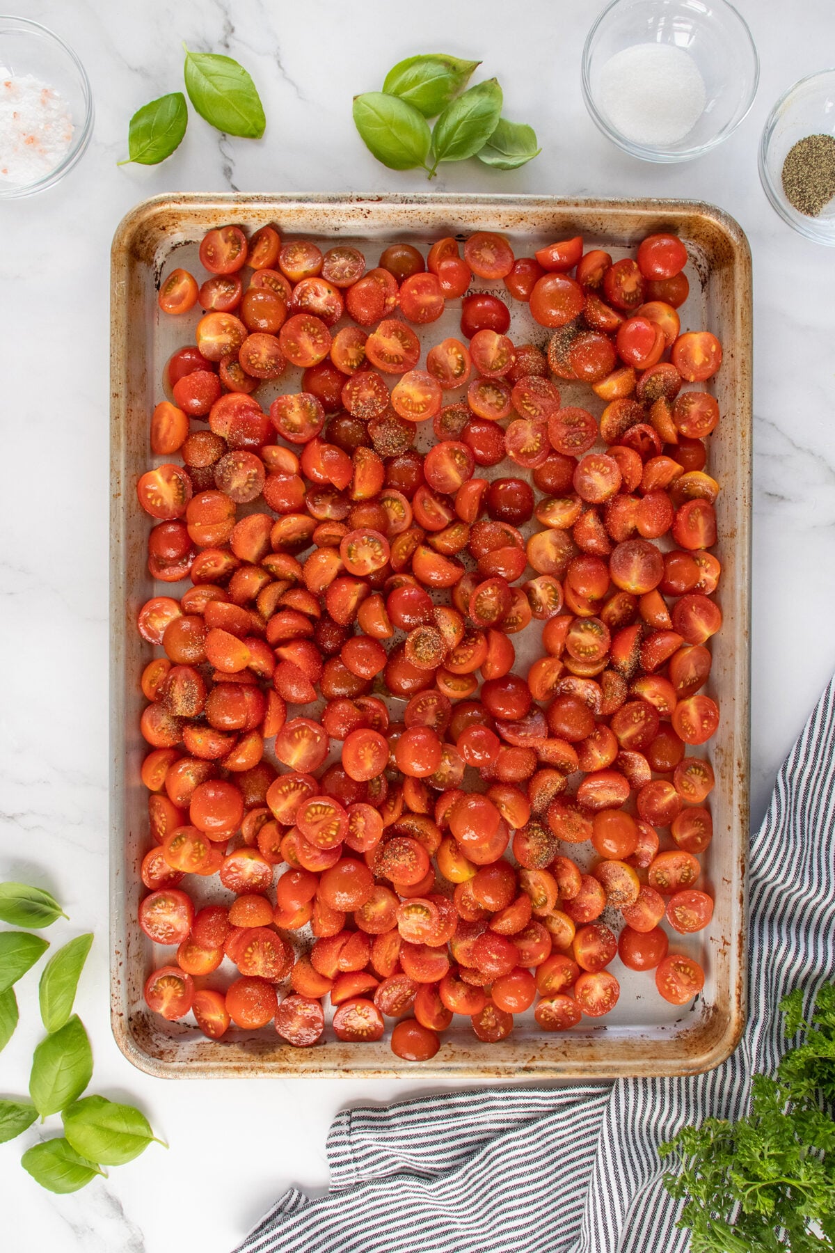 A baking sheet filled with halved cherry tomatoes, sprinkled with seasoning, sits on a white surface. Fresh basil leaves, a striped towel, and small bowls with spices surround the tray.