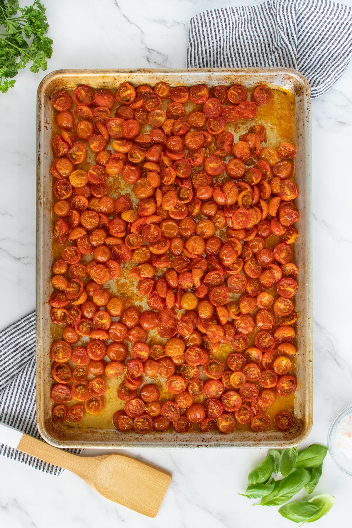 A baking sheet filled with roasted cherry tomato halves, surrounded by a wooden spatula, fresh basil, parsley, and striped kitchen towels on a marble countertop.