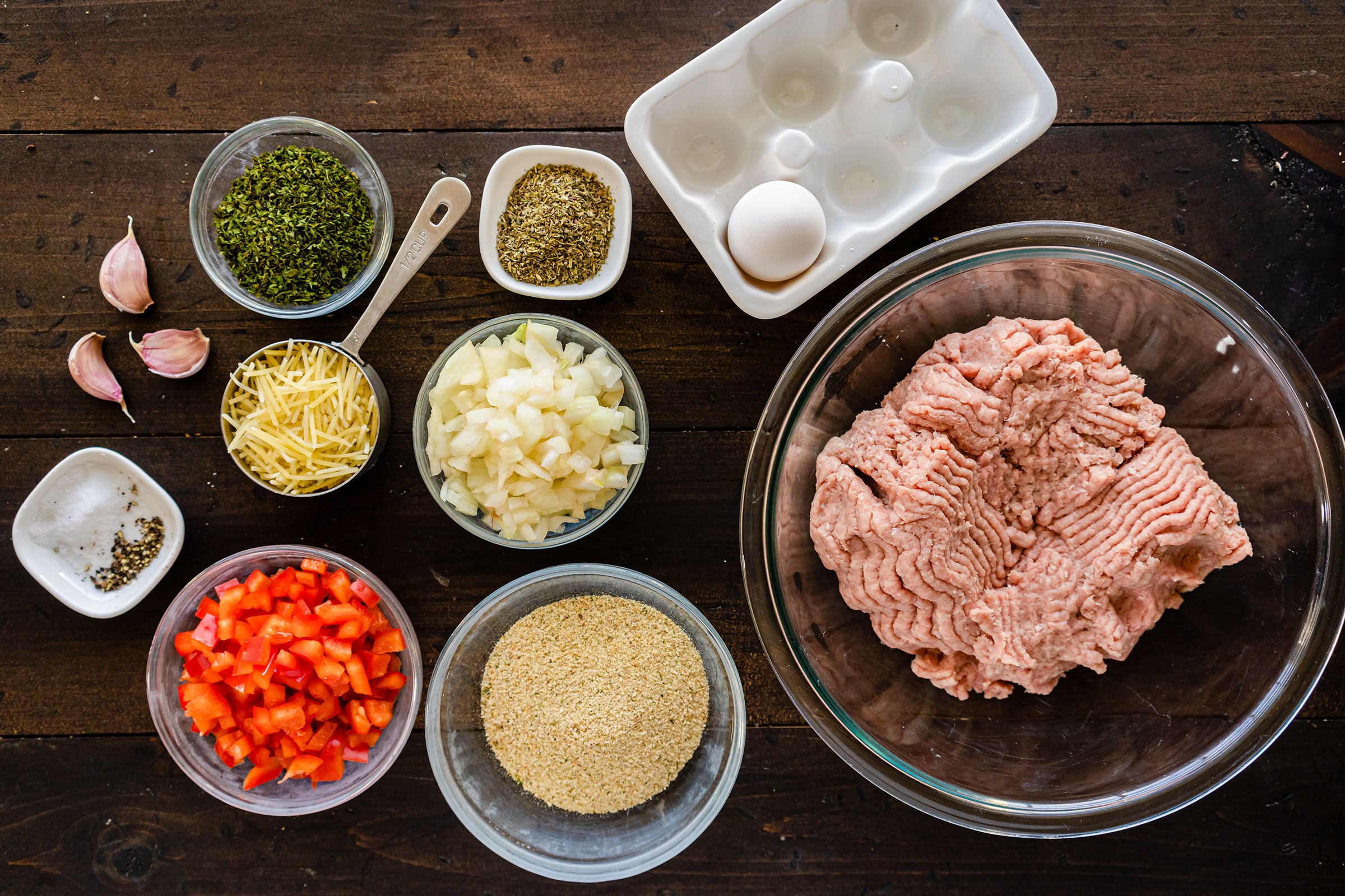 Ingredients arranged on a wooden table, including ground meat, breadcrumbs, chopped onion, diced red pepper, shredded cheese, herbs, spices, garlic, an egg, and an empty egg carton.
