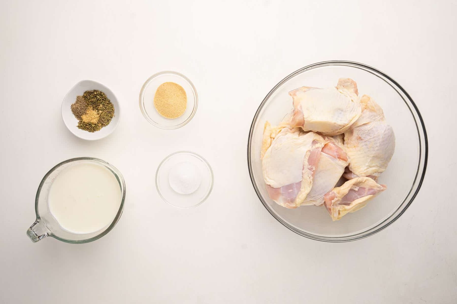 A glass bowl of raw chicken pieces, a measuring cup of buttermilk, a small bowl of salt, a bowl of garlic powder, and a bowl with mixed dried herbs on a white surface arranged for cooking prep.
