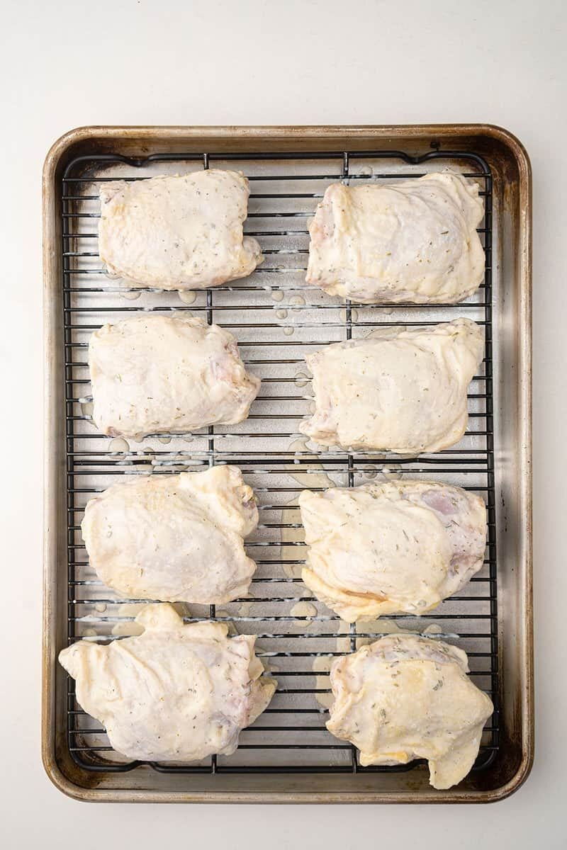 Eight raw, seasoned chicken thighs are arranged on a wire rack set over a baking sheet, ready for cooking. The coating on the chicken pieces appears creamy and lightly peppered.