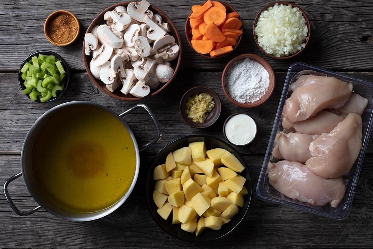 Overhead view of soup ingredients on a wooden table: raw chicken breasts, diced potatoes, chicken broth in a pot, sliced mushrooms, carrots, celery, chopped onions, minced garlic, flour, cream, and spices in bowls.