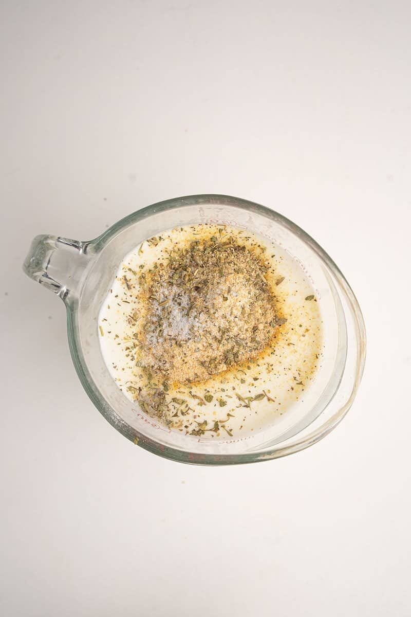 A glass measuring cup filled with a creamy liquid, topped with a pile of dried herbs and ground spices, viewed from above on a white background.