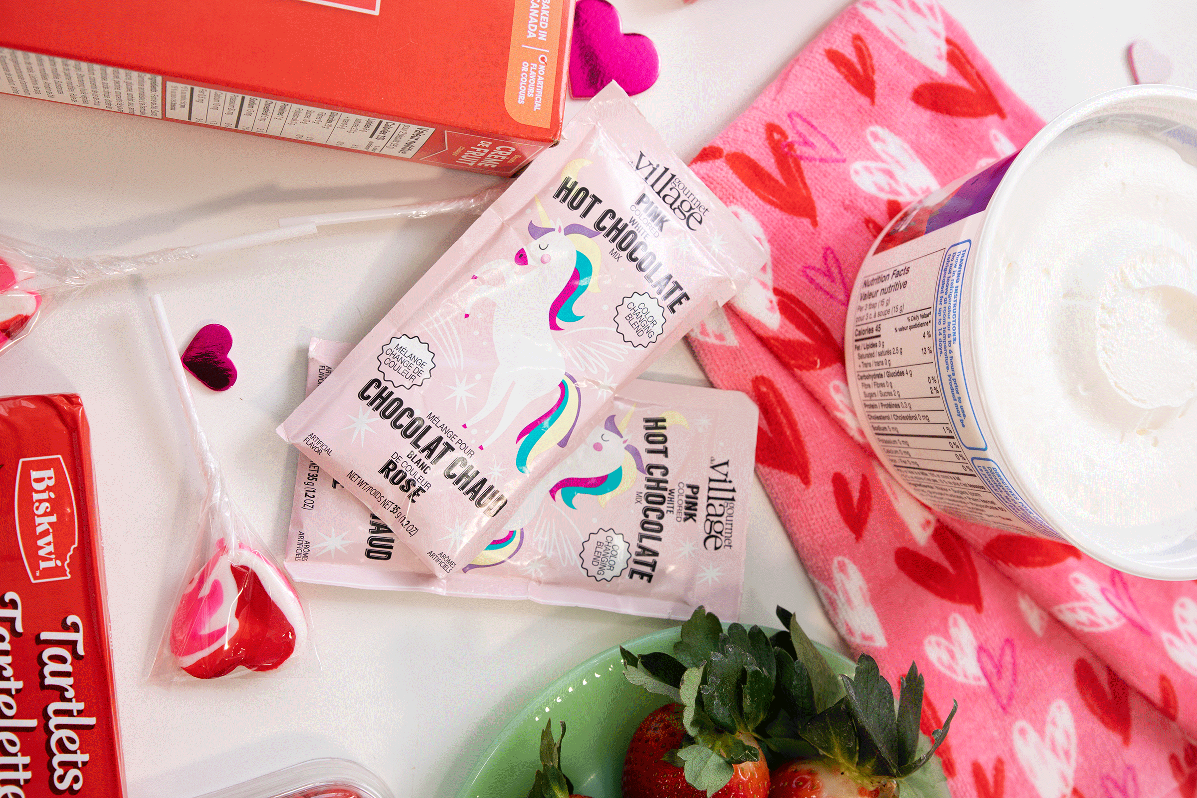 Packets of unicorn-themed rose hot chocolate, strawberries, whipped topping, cookies, a box of tartlets, and a pink heart-patterned napkin are arranged on a white surface with small pink heart decorations.