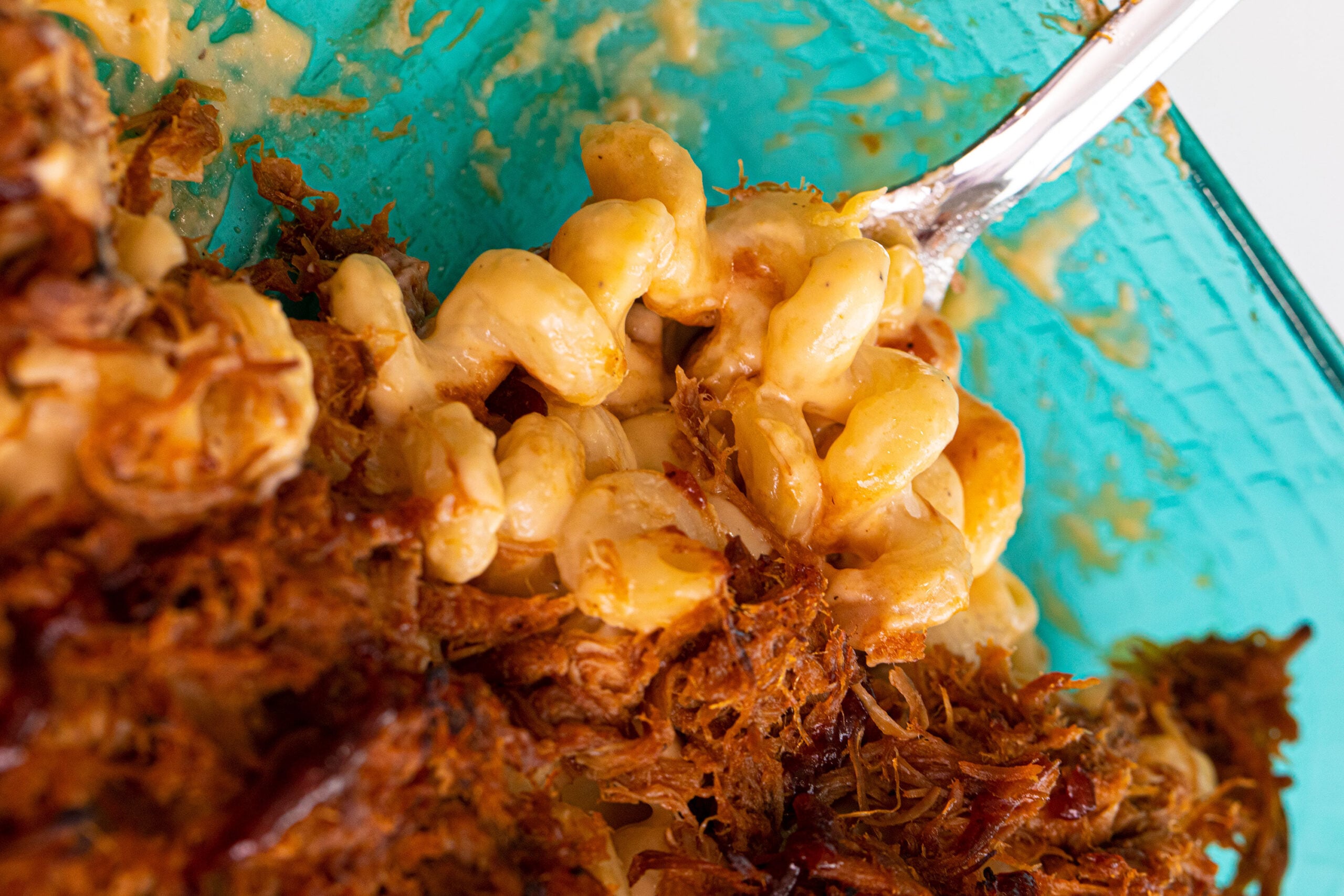 Close-up of creamy macaroni and cheese in a glass dish, mixed with shredded pulled pork. A fork is lifting a portion of the cheesy pasta and pork mixture.