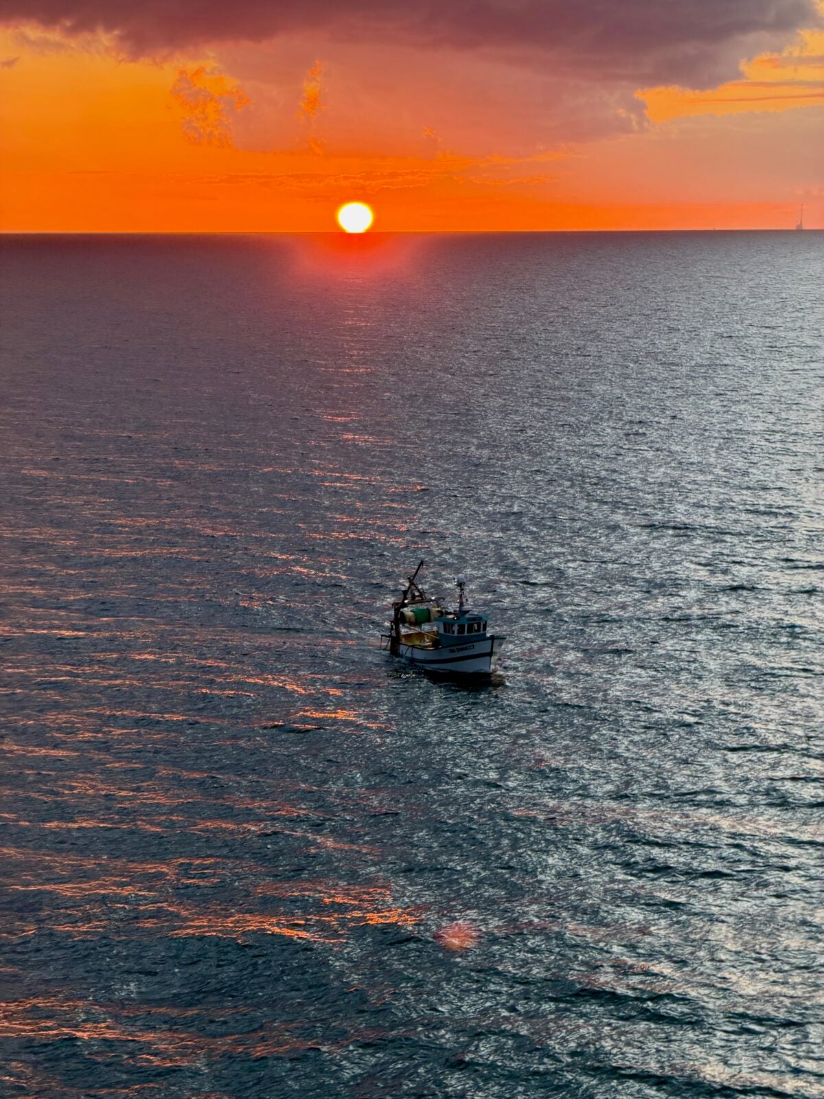A small fishing boat floats on calm water under a vibrant orange sunset, with the sun low on the horizon and clouds tinged with warm colors above the open sea.