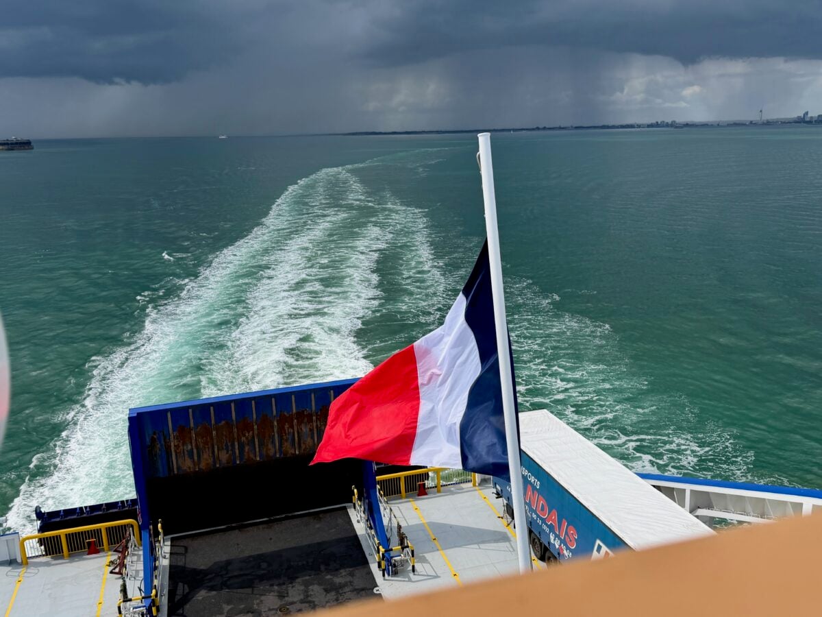 A French flag waves at the stern of a ferry moving across greenish-blue water, leaving a white wake behind. Dark clouds loom over the distant shoreline under a moody sky.
