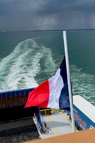A French flag waves at the stern of a ferry moving across greenish-blue water, leaving a white wake behind. Dark clouds loom over the distant shoreline under a moody sky.
