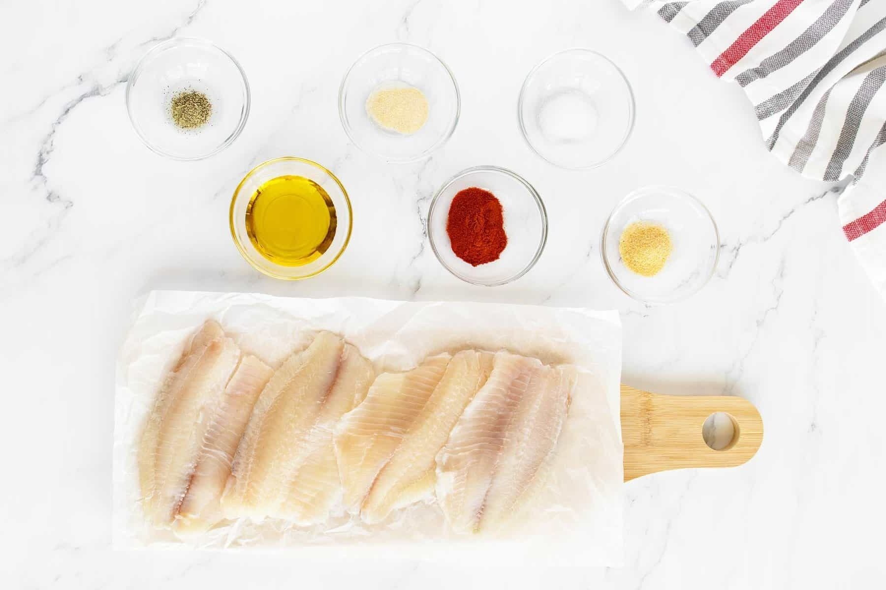 Five raw fish fillets on parchment paper atop a wooden cutting board, surrounded by small glass bowls containing seasoning powders, oil, salt, pepper, and a striped kitchen towel on a white surface.