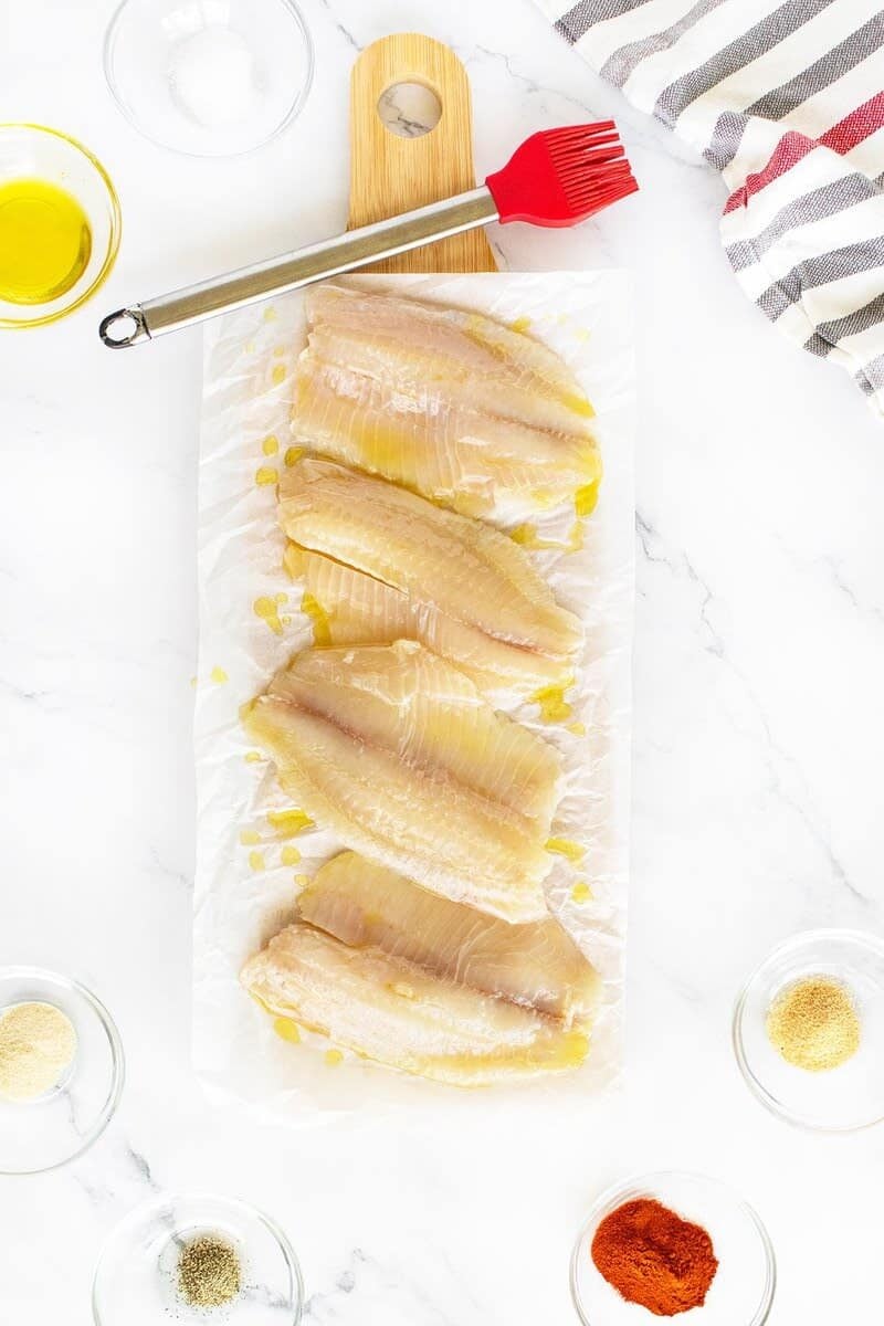 Four raw fish fillets are laid out on parchment paper on a wooden board, drizzled with oil. Surrounding them are seasonings in small bowls, a red basting brush, and a striped kitchen towel.