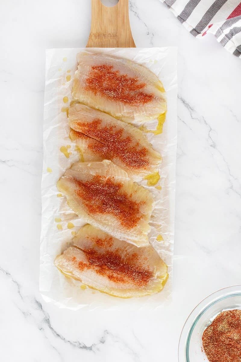 Four raw fish fillets on parchment paper with a wooden handle, seasoned with paprika and spices, arranged on a white marble surface. A striped cloth and a small bowl of seasoning are nearby.