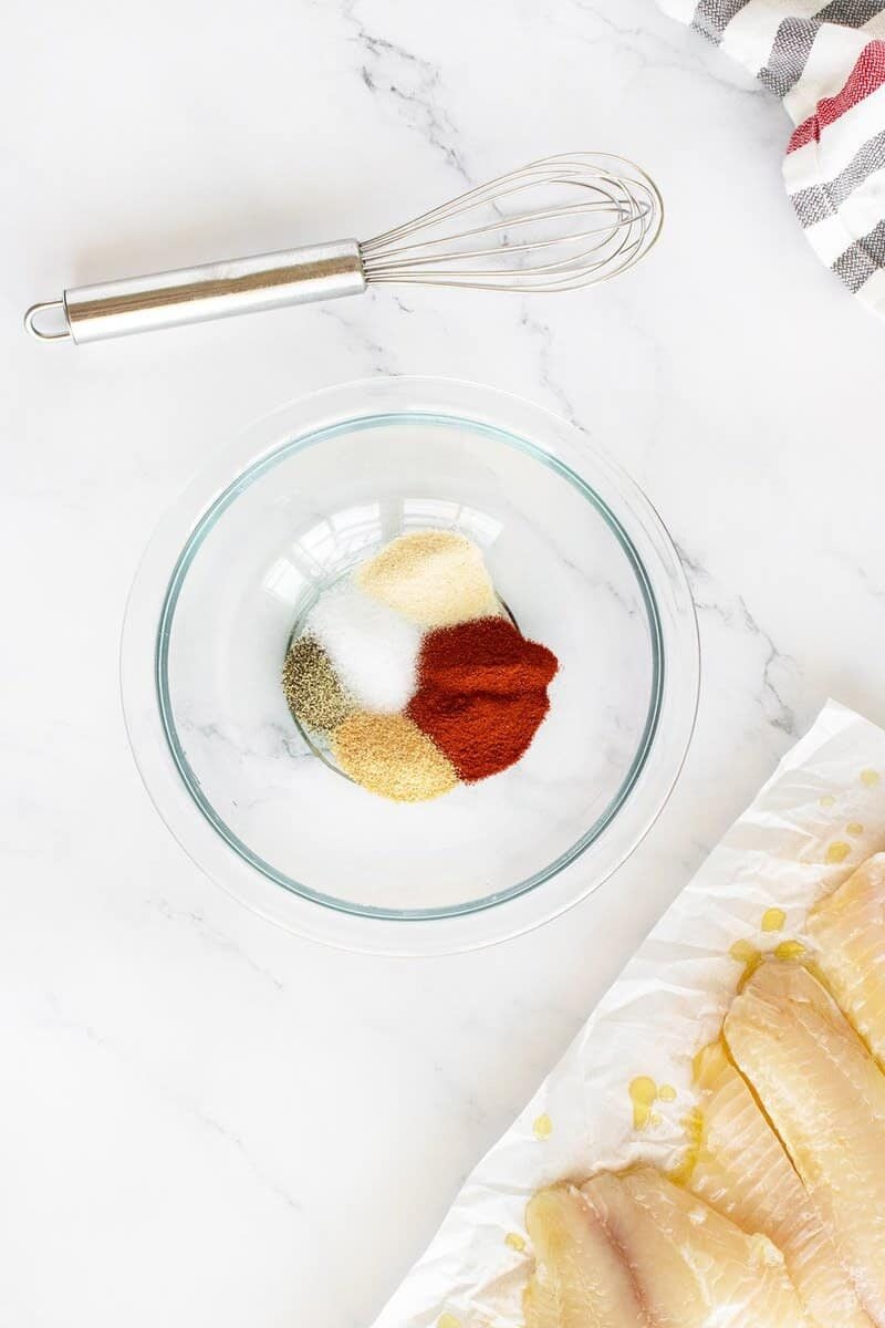 Clear glass bowl with piles of different spices, including paprika, onion powder, garlic powder, and salt, next to raw fish fillets on parchment paper and a metal whisk on a white marble countertop.
