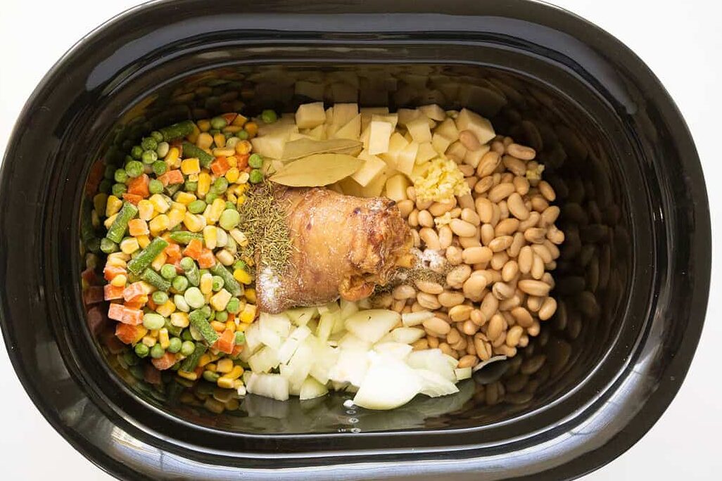 A slow cooker filled with mixed vegetables, diced potatoes, white beans, chopped onions, bay leaves, garlic, herbs, and a ham hock, all arranged in separate sections before cooking.