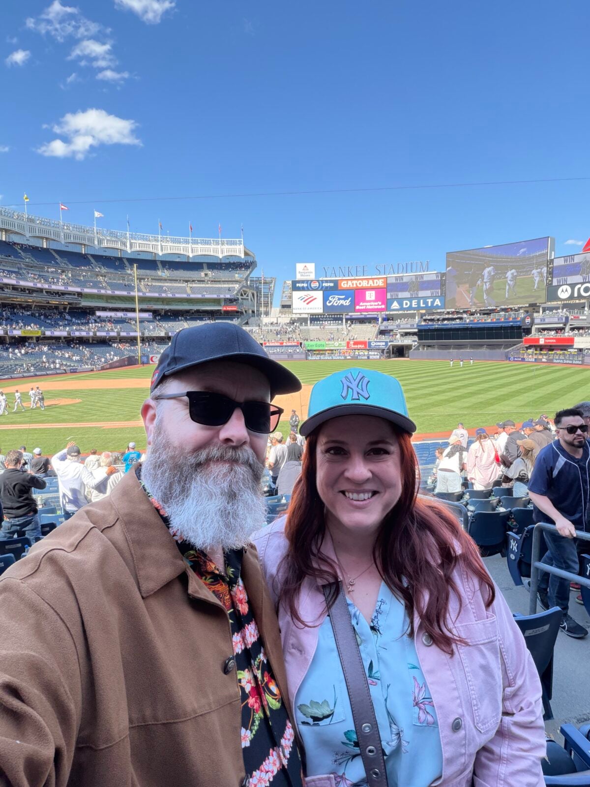 A man and woman smiling for a selfie at a baseball stadium with the field, scoreboard, and crowd visible in the background on a sunny day. The woman wears a teal New York Yankees cap.