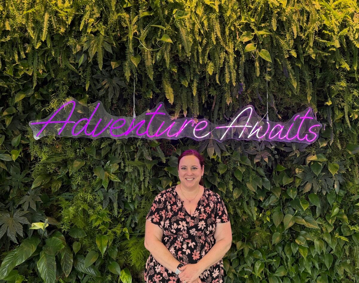 A person in a black floral dress stands smiling in front of a lush green plant wall with a neon sign that reads Adventure Awaits.