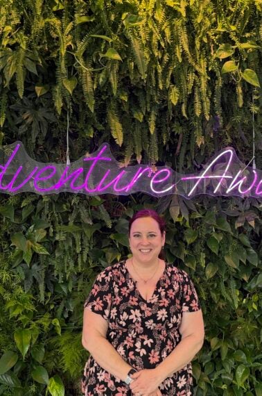 A person in a black floral dress stands smiling in front of a lush green plant wall with a neon sign that reads Adventure Awaits.