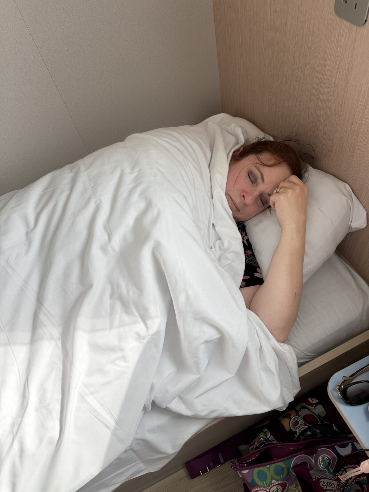 A person with red hair is sleeping on a bed, covered with a white blanket. They are resting their head on a pillow, with one hand near their face. The room has light-colored walls and bedding.