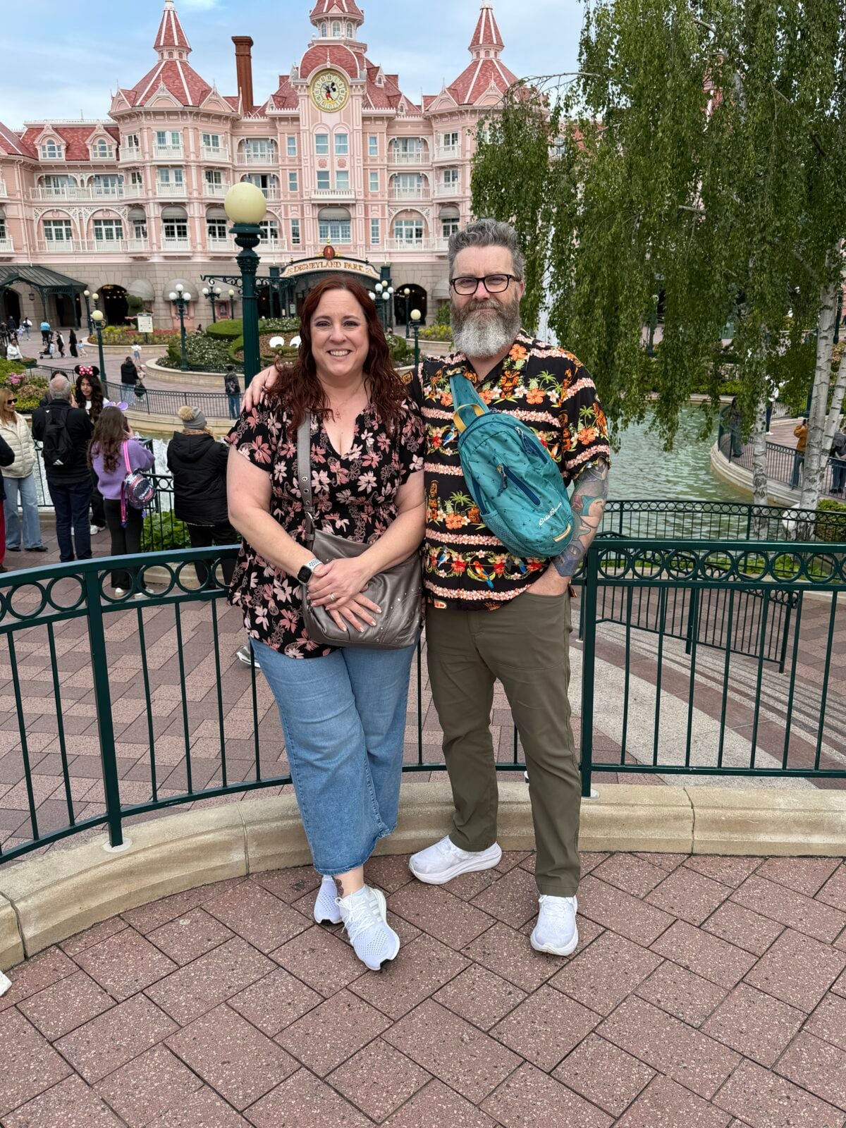 A smiling couple poses in front of a pink, ornate building with a clock tower, surrounded by greenery and water features. Both wear casual clothes and stand next to a black metal fence. People are in the background.