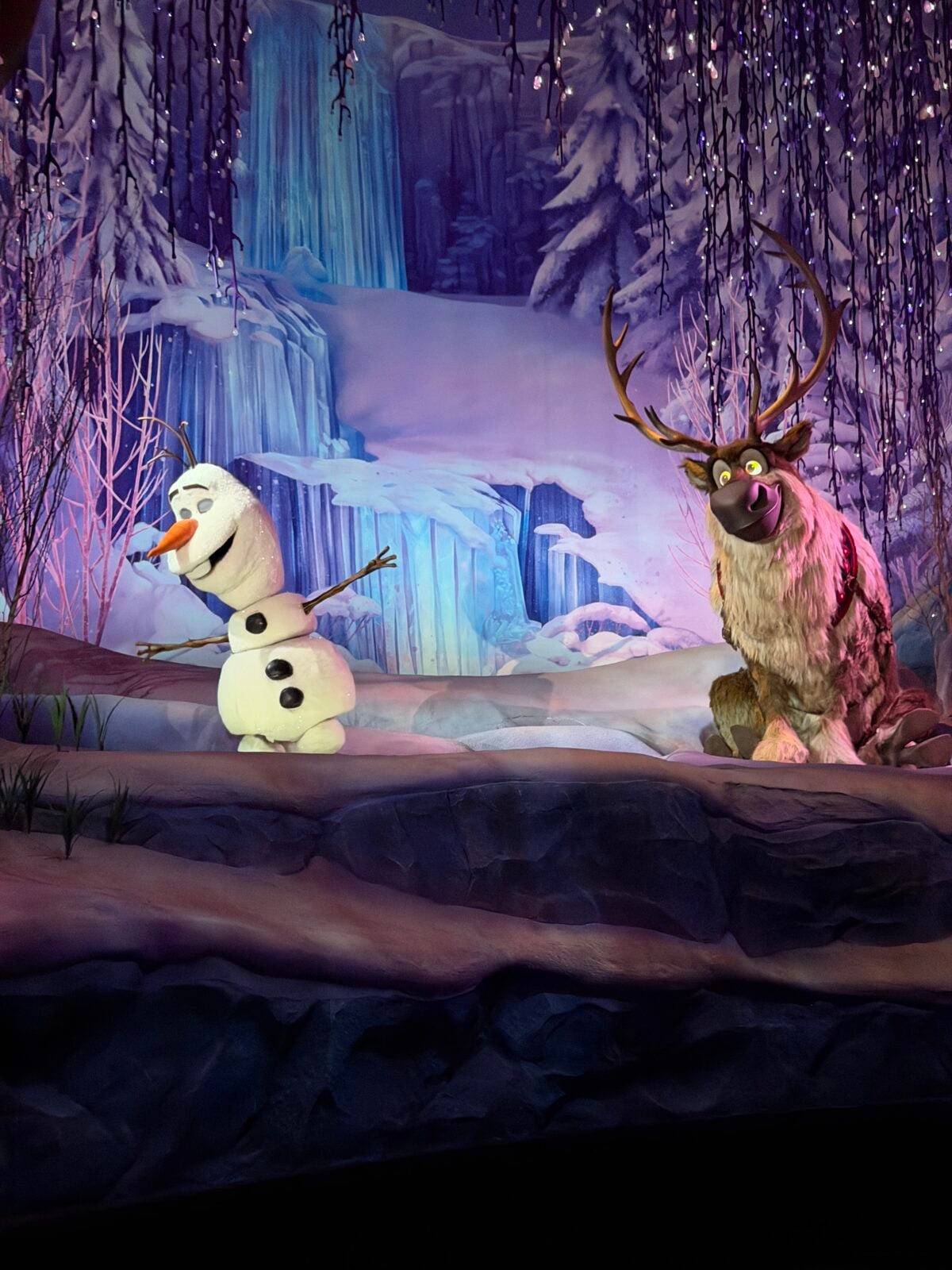 Animatronic figures of Olaf the snowman and Sven the reindeer from Frozen are set against a snowy, winter backdrop with icy blue waterfalls and frosted trees. Purple lighting creates a magical atmosphere.