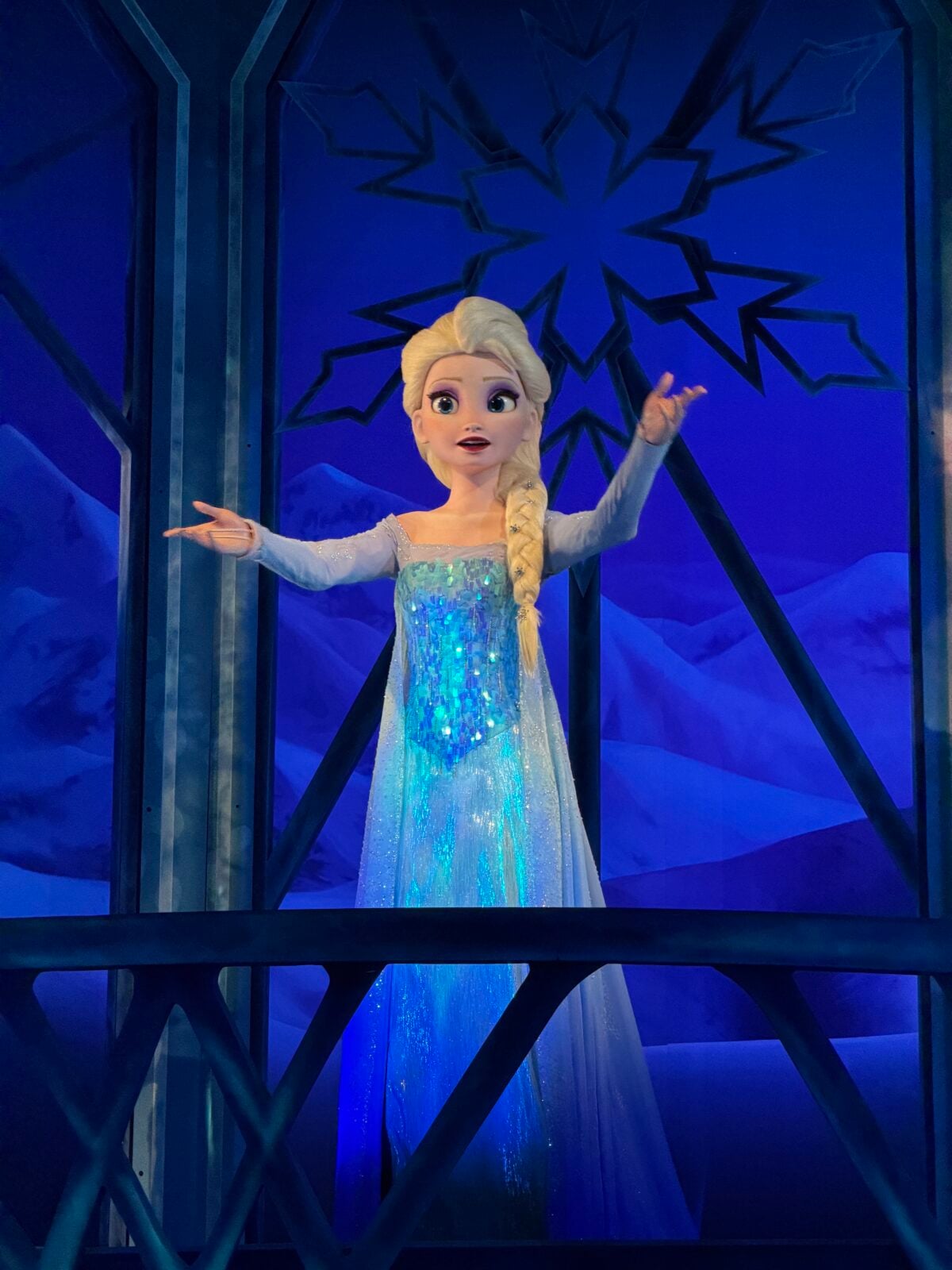 A lifelike Elsa animatronic figure from Disney’s Frozen stands on a balcony, arms outstretched, wearing a sparkling blue dress with a snowy backdrop and snowflake design behind her.