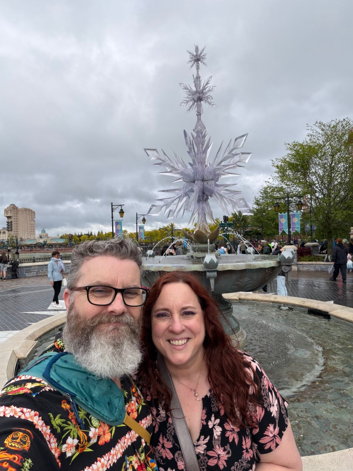 A smiling man with glasses and a woman with red hair pose in front of a fountain featuring a large, decorative ice crystal structure in an outdoor park on a cloudy day. People and trees are visible in the background.