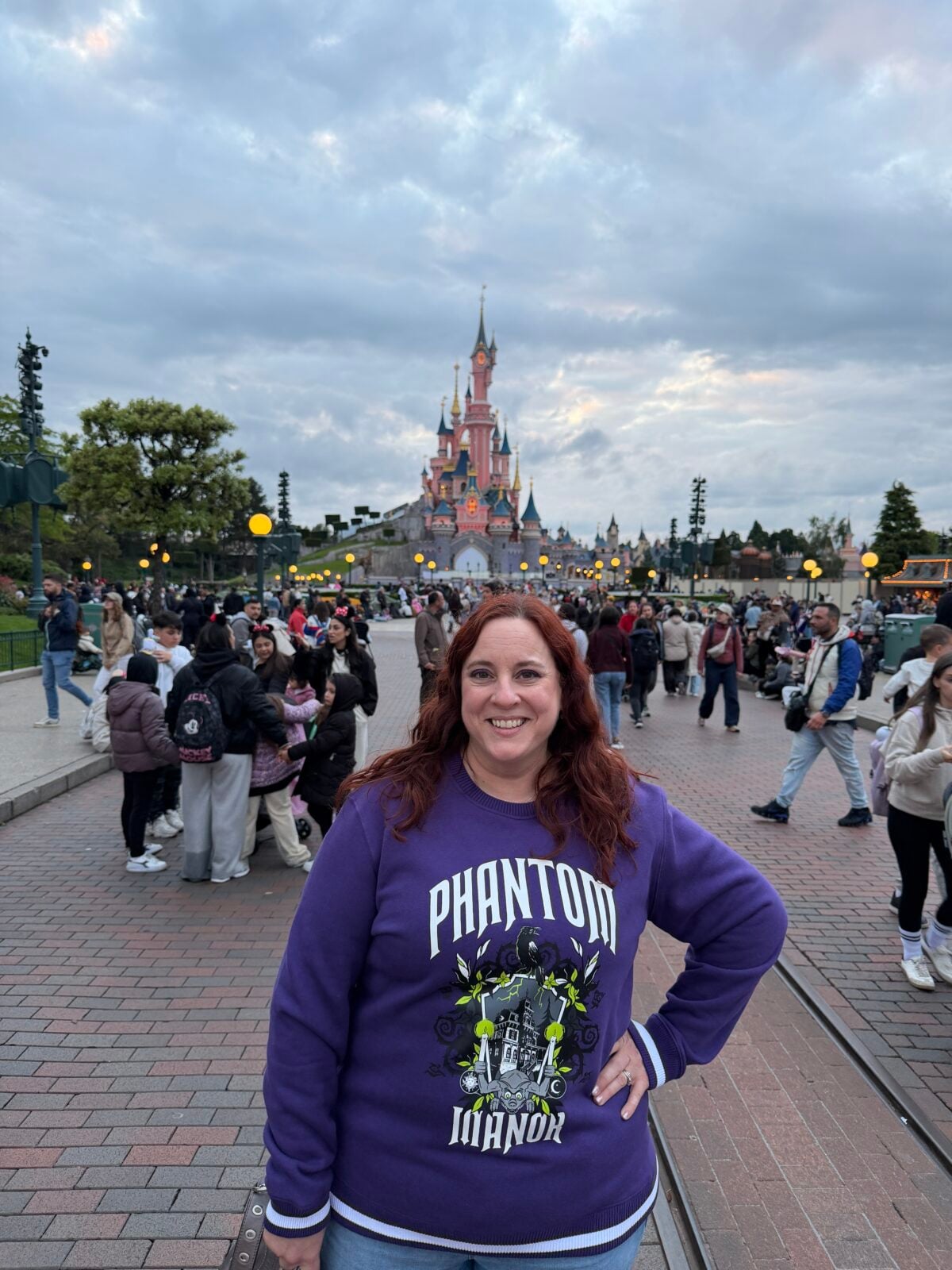 A woman with red hair smiles at the camera in front of Disneyland’s Sleeping Beauty Castle, wearing a purple Phantom Manor sweatshirt. Crowds and colorful lights surround the castle under a cloudy sky.