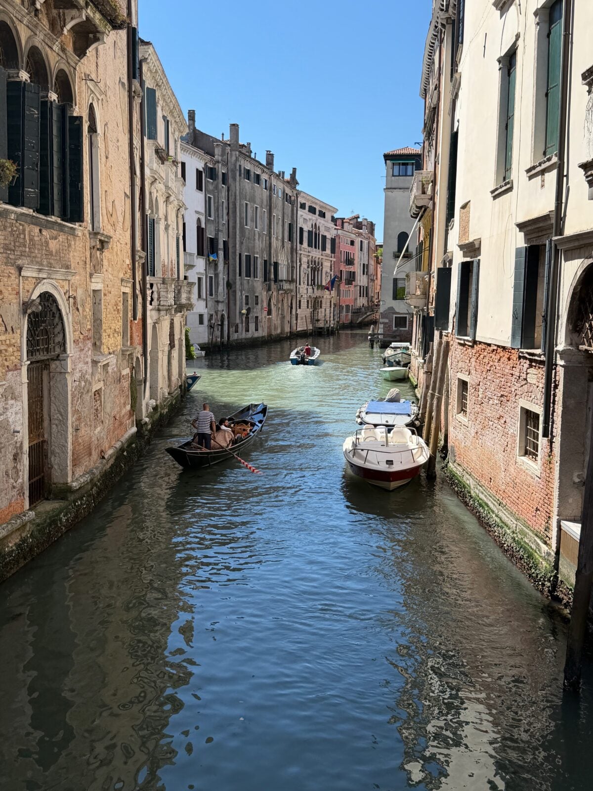 A narrow canal in Venice with old buildings on both sides, small boats docked along the walls, and a man rowing a classic gondola in Venice under a clear blue sky.