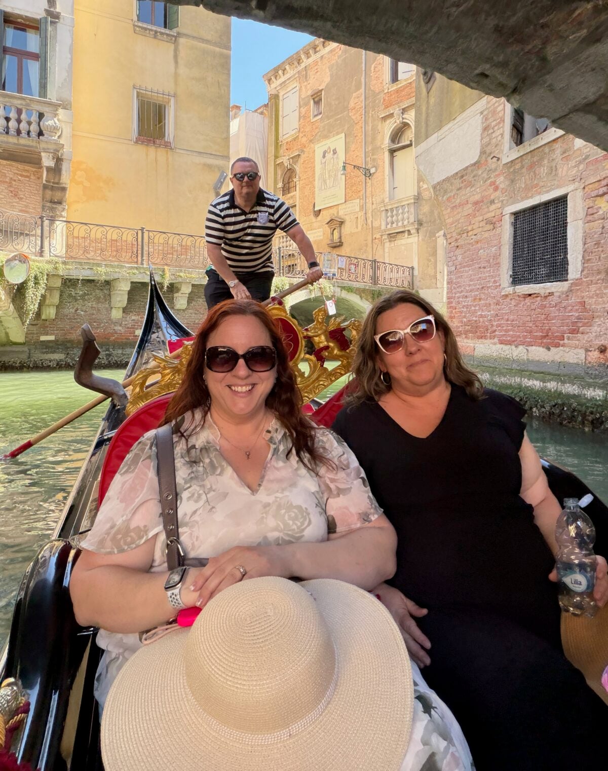 Two women sit in a gondola smiling, one holding a sunhat, as a gondolier in a striped shirt stands behind them steering through a canal in Venice, with historic buildings and a footbridge in the background.