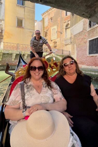 Two women sit in a gondola smiling, one holding a sunhat, as a gondolier in a striped shirt stands behind them steering through a canal in Venice, with historic buildings and a footbridge in the background.