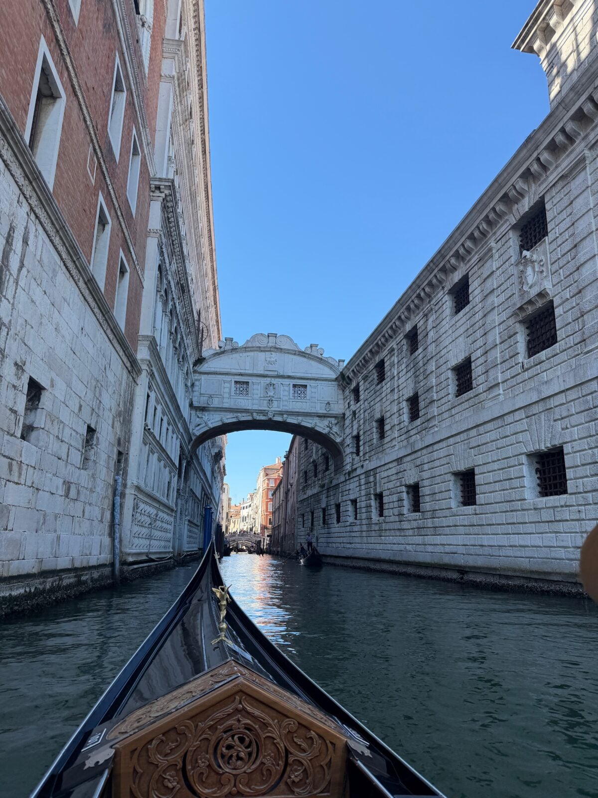 A view from a gondola on a canal in Venice, looking towards the historic white Bridge of Sighs connecting two stone buildings under a clear blue sky.