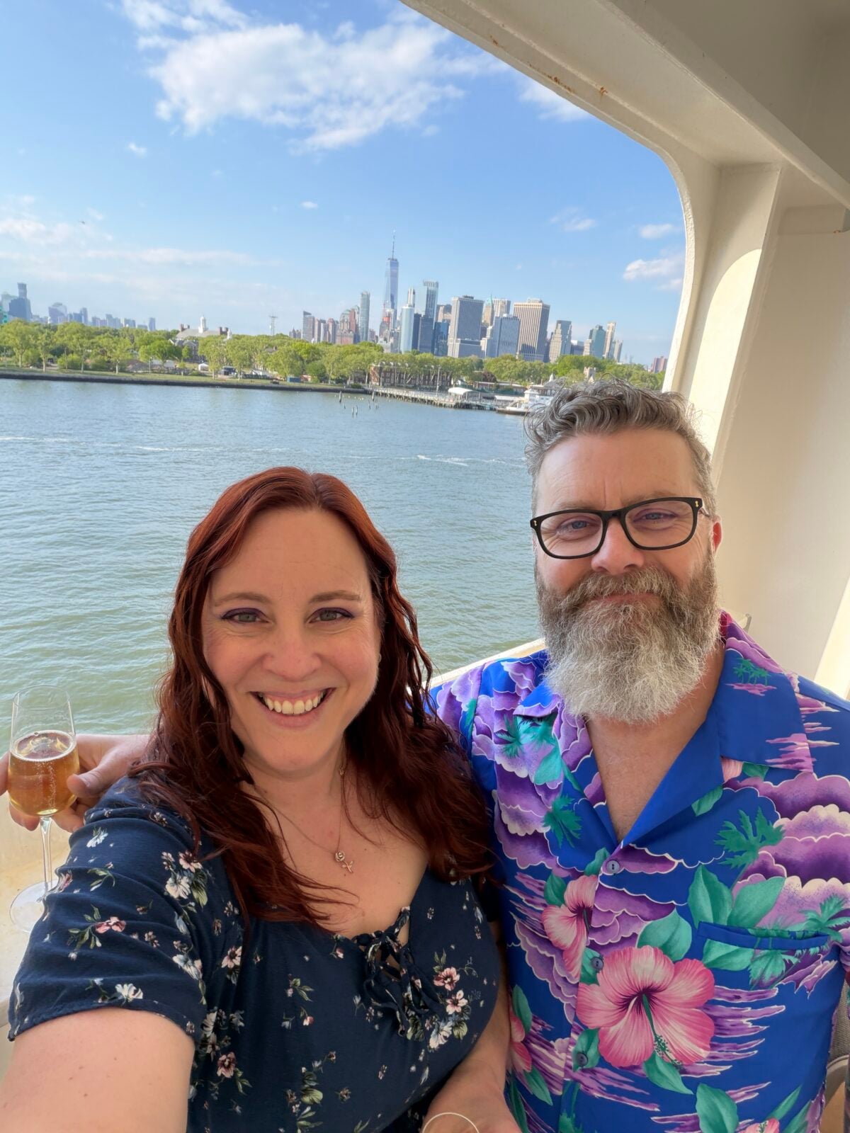 A smiling couple takes a selfie on a boat with the New York City skyline in the background under a blue sky. The woman has long red hair and wears a dark floral dress; the man has gray hair, a beard, and a blue floral shirt.