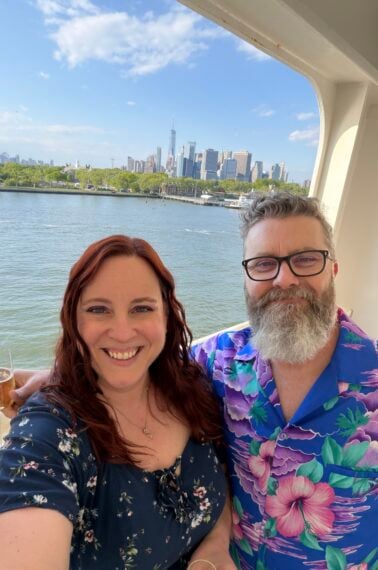 A smiling couple takes a selfie on a boat with the New York City skyline in the background under a blue sky. The woman has long red hair and wears a dark floral dress; the man has gray hair, a beard, and a blue floral shirt.